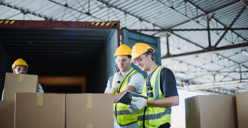 Logistics workers in safety vests and helmets loading packages from container truck, using tablet for inventory check. Industrial teamwork scene in spacious cargo warehouse.