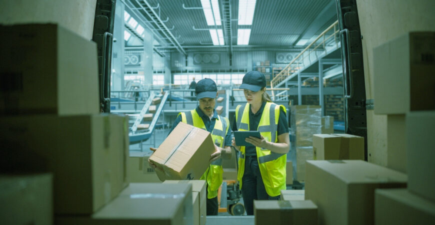 Caucasian Female Stocking Associate Using Tablet Computer To Check Inventory In Logistics Facility With Conveyor Belt. Male Warehouse Worker Loading Boxes In Van For Delivering Orders To Clients.